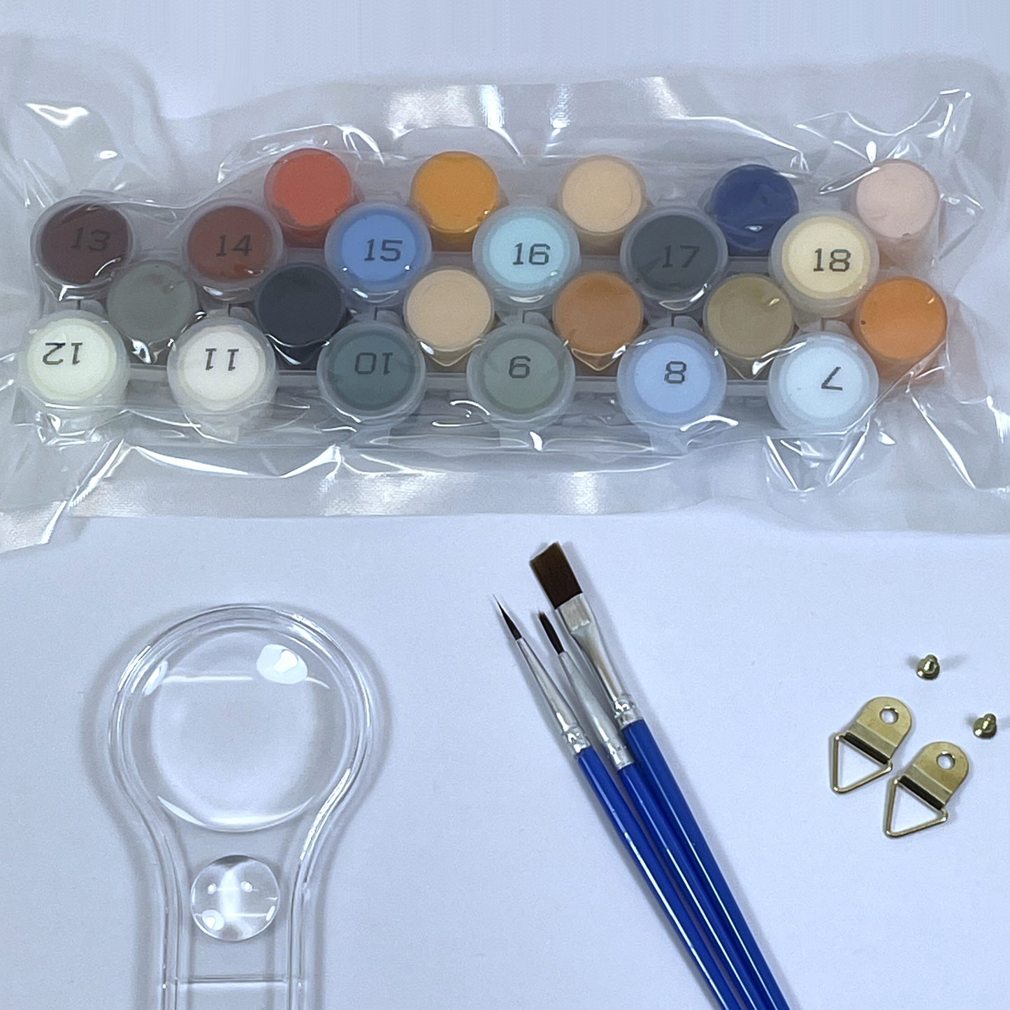 Set of paint pots, three paint brushes and a magnifying glass on a white surface.